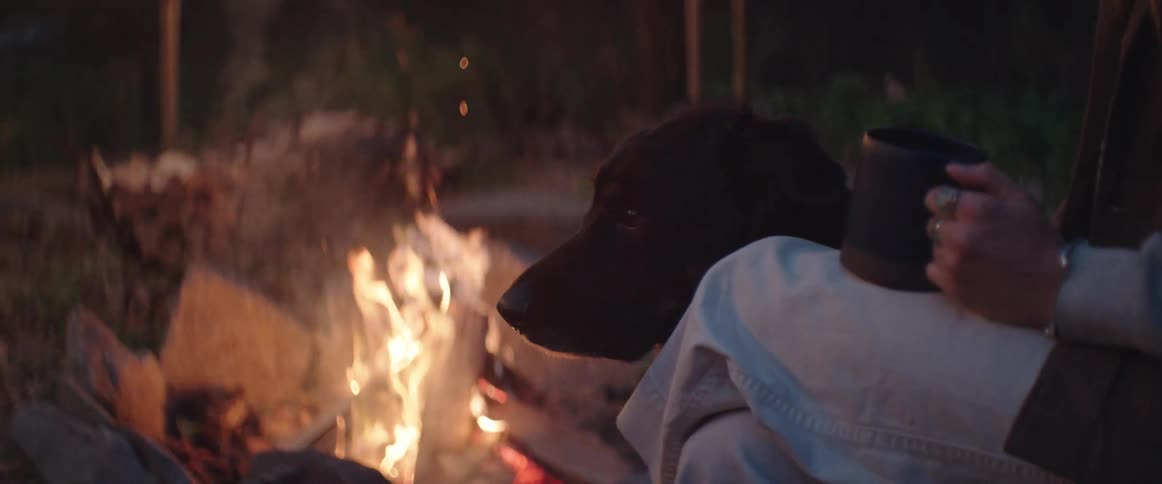A woman holds a mug and sits by a fire blazing in a firepit with her dog