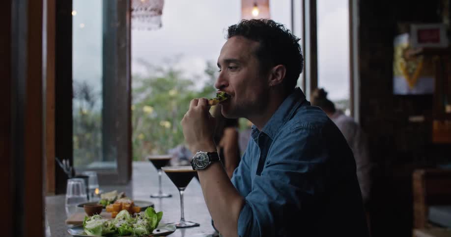 A man sits in a restaurant with a cocktail and eats pizza and looks at his phone and sets it down