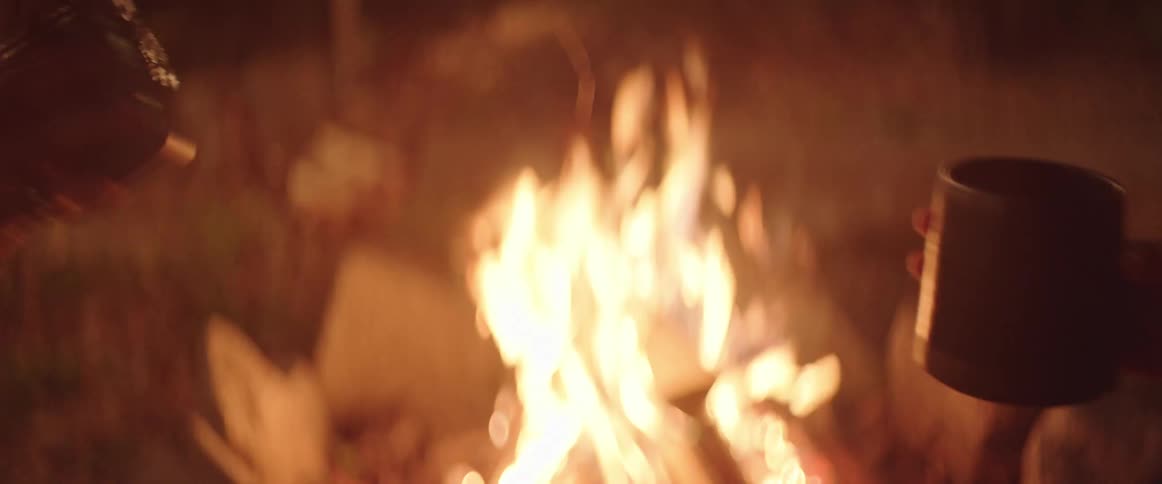 A man and woman clink mugs over a campfire
