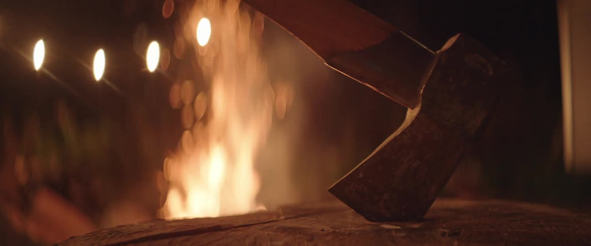 An axe sits in a log in front of a campfire