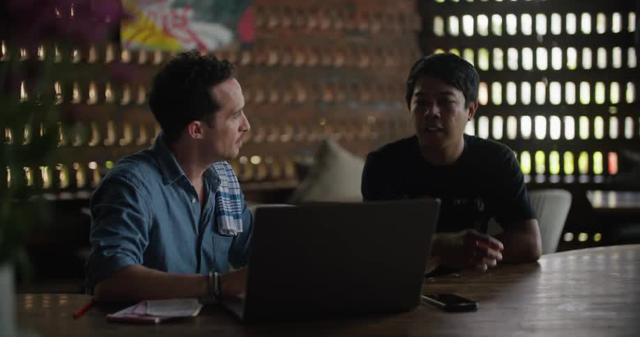 A pair of men speak while sitting in front of a laptop at a counter