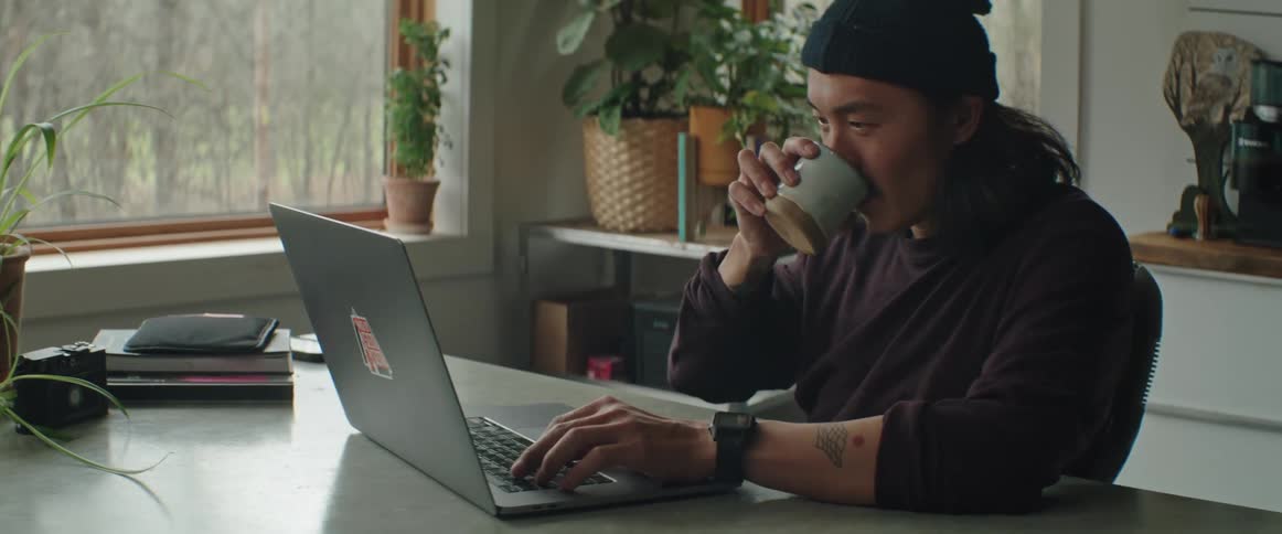 A man sits at home and drinks coffee as he looks at his laptop