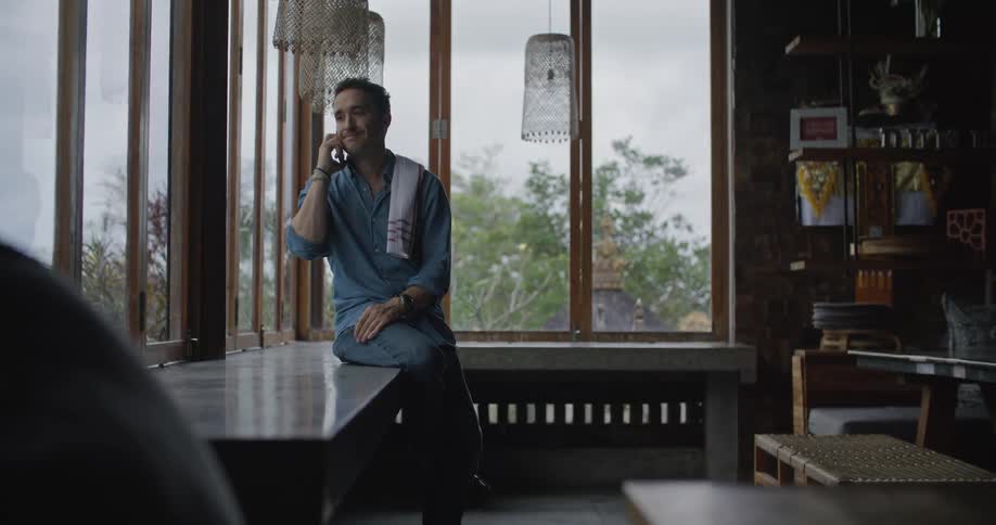 A man sits on a table next to a large window while talking on his phone