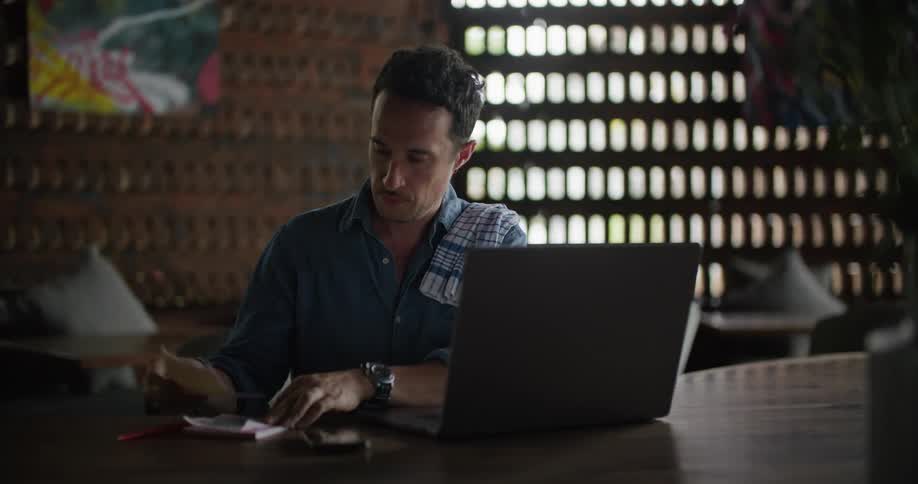 A man carries a notepad to his laptop and sits down before writing while referencing the screen