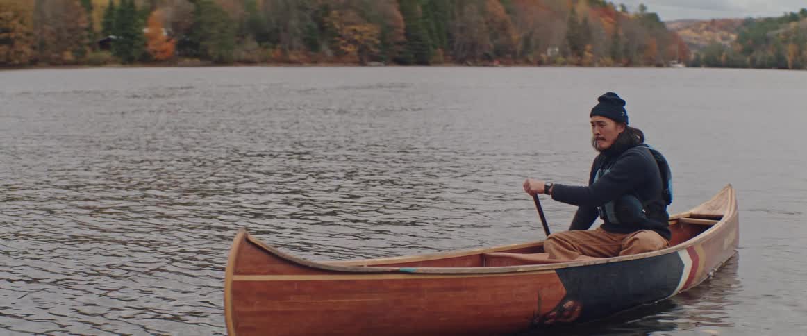 A man is rowing a canoe as he looks around and there are mountains covered with trees in the distance