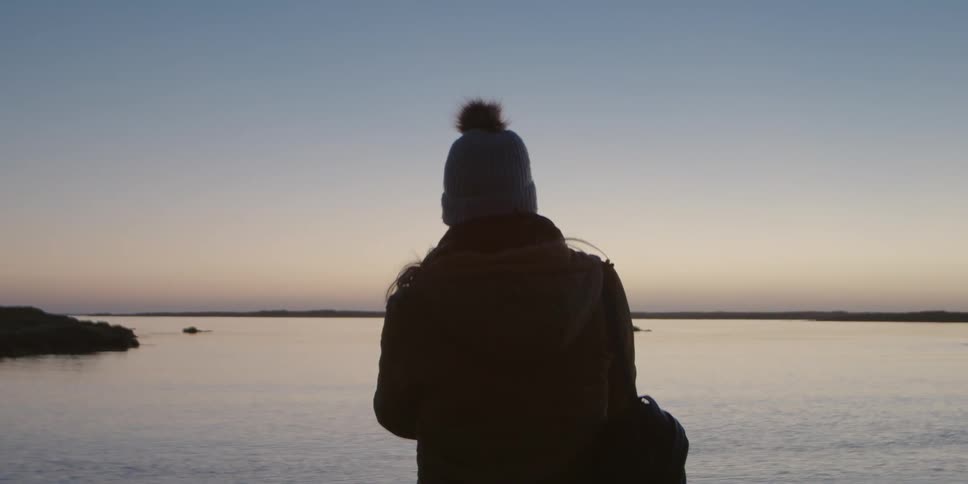 Person wearing jacket and knit hat stands on shore looking at water