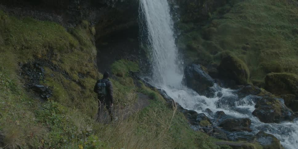 A hiker looks around at his surroundings while walking along a narrow dirt trail towards a waterfall