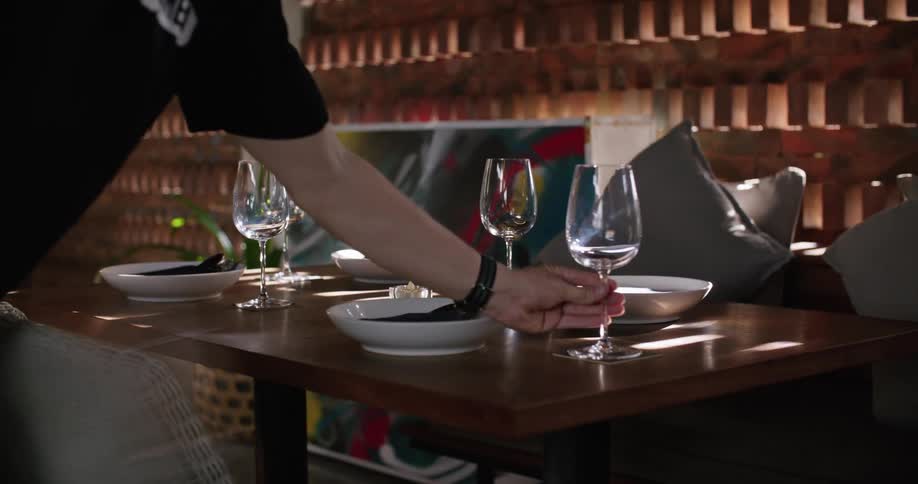 A person sets a large wine goblet down on a dining table that is being set