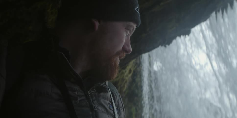 A man stands under a cliff as a waterfall cascades down it
