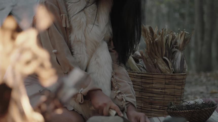 A native american woman is sitting next to a teepee cutting up something to cook on the fire