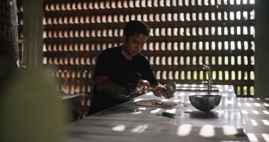 A male restaurant worker spooning sauce onto a pizza crust in the kitchen