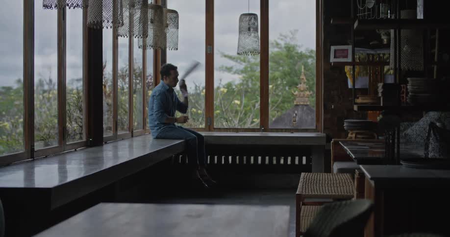 A man is sitting on a counter near windows folding a towel and he throws it over his shoulder