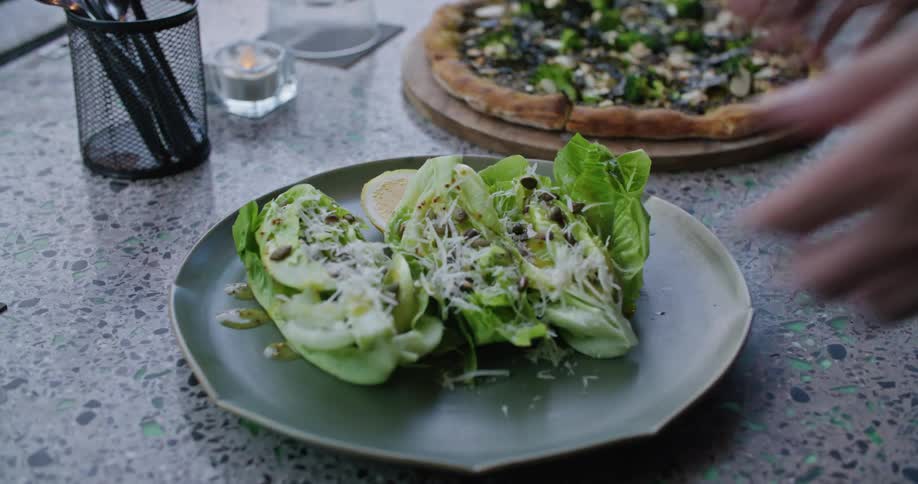 Person places plated salad on stone counter next to pizza