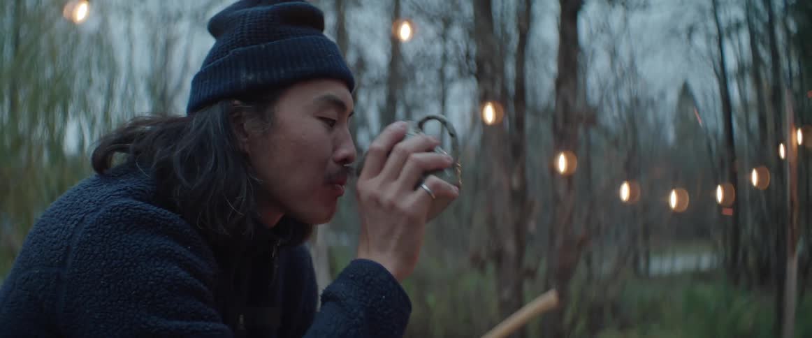 Man with long hair drinks from a cup in front of a campfire at evening