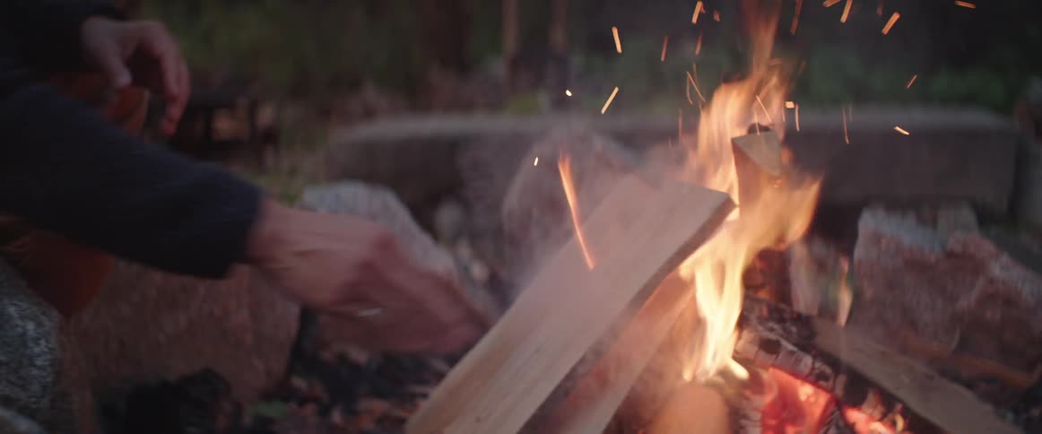 Flames rise as a man puts wood on a camp fire