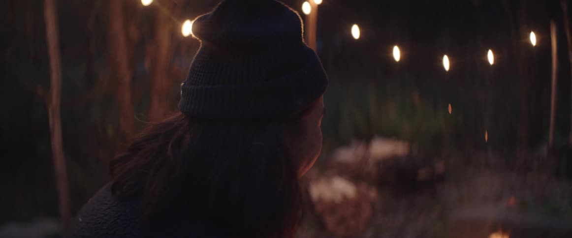 Man laughs while sitting near lit campfire in area strung with lights