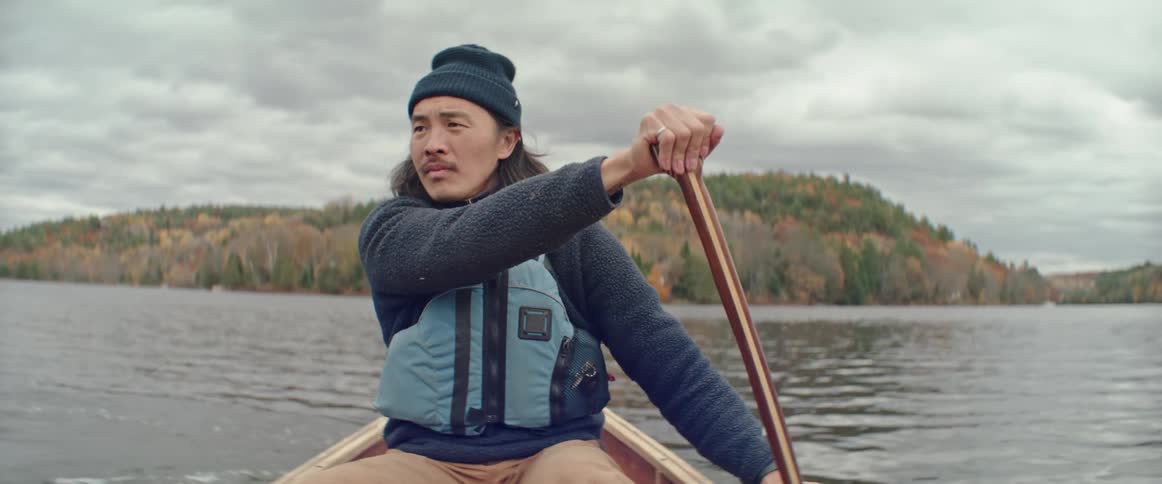 A man is rowing a canoe as he looks around and there are mountains covered with trees in the distance