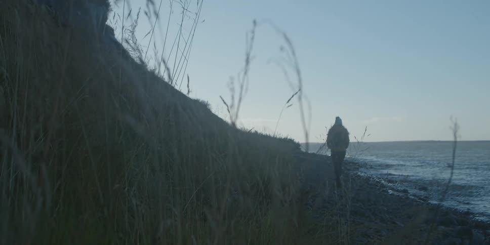 A person wearing a coat and hat is walking along the shoreline with their hands in their pockets as the waves roll in