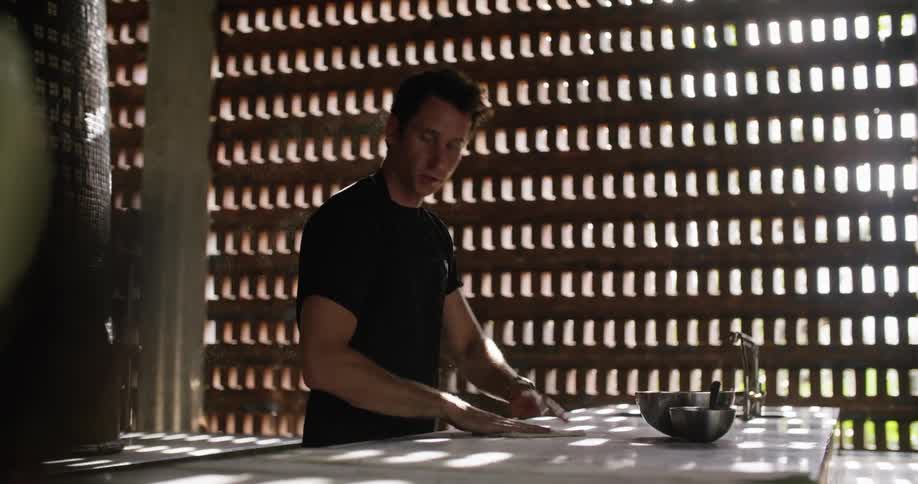 Man smiles while working with dough in restaurant kitchen near wall designed with small openings to let the sun shine through