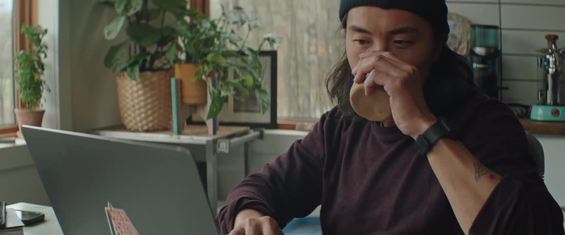 Man with long hair and mustache drinks coffee while working on a laptop at home