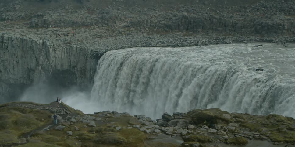 A few people stand at a cliff to observe a large and powerful waterfall