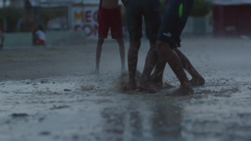 A group of boys play soccer under a sprinkler in a muddy field