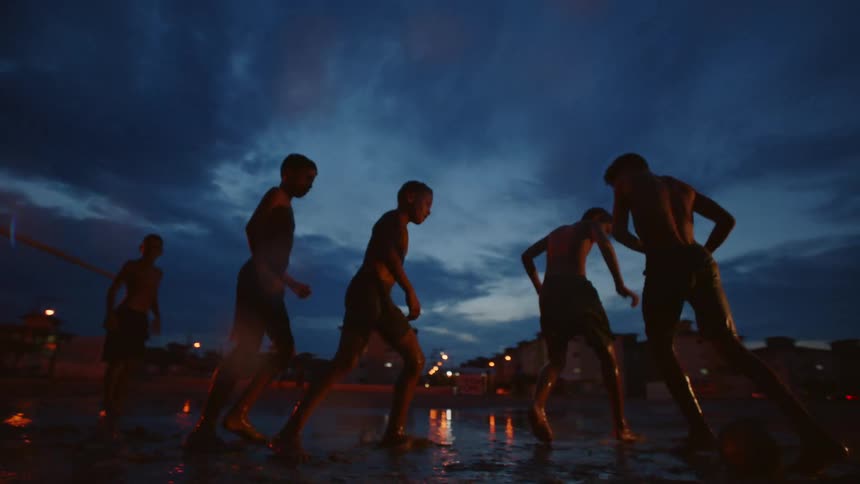 A group of boys are splashing in the water and mud as they play soccer in puddles of water in the rain