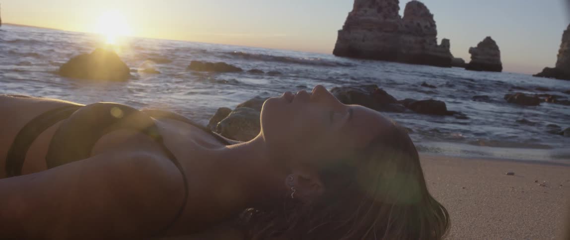 A woman lays on the sand at the water's edge on a beach at dusk