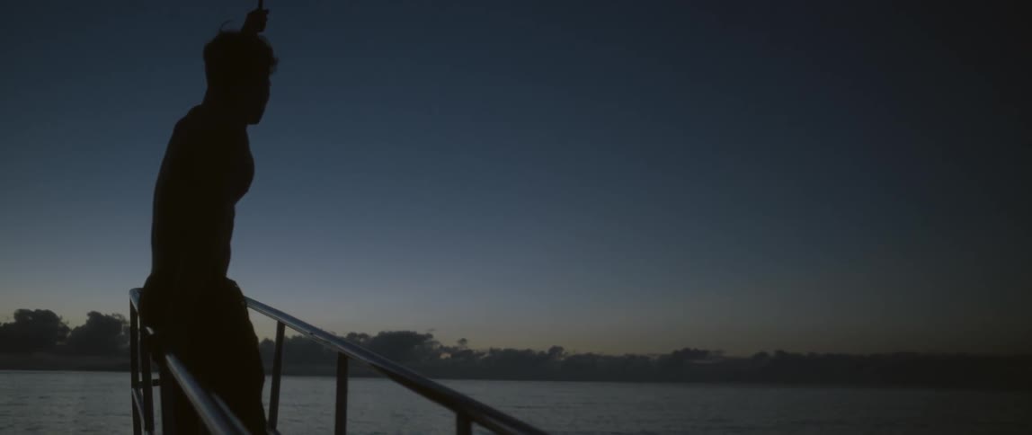 A man holding on to a rope as he stands on a boat deck while night falls