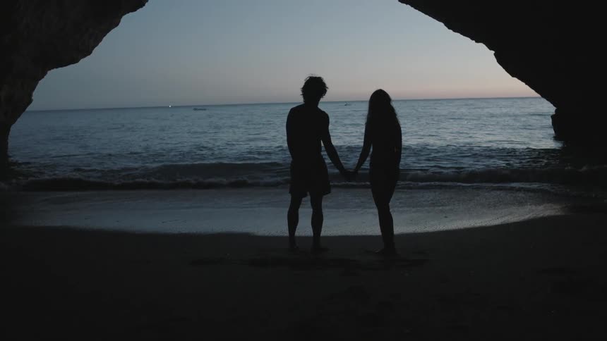 A man and woman stand on the beach near a natural arch holding hands at dusk