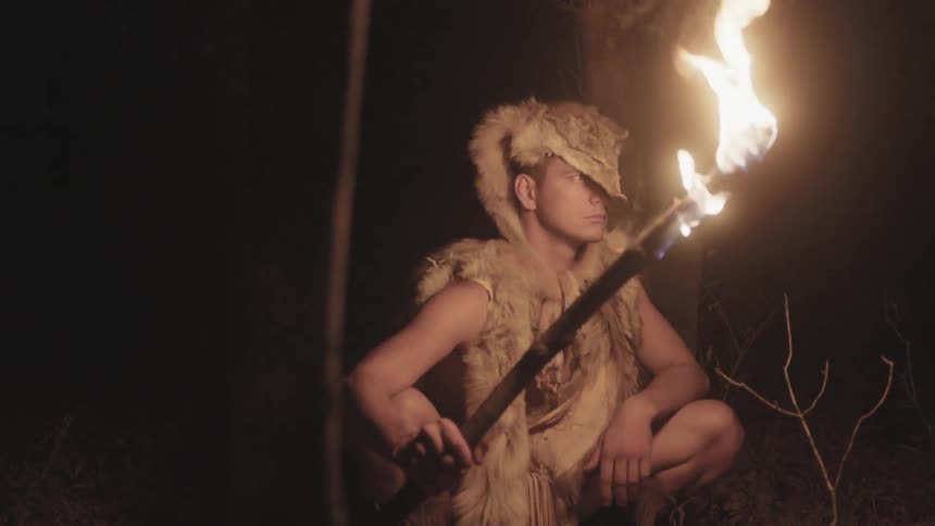 A man holding a torch and wearing animal fur clothing is crouched in a forest at night