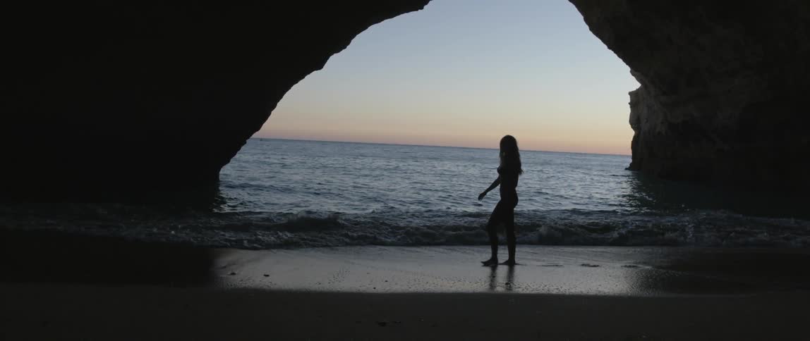 A woman walks at the water's edge near an arch on a beach at sunset