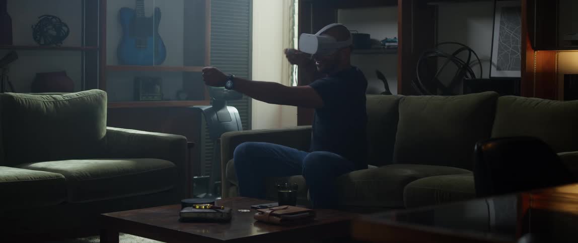 A man is sitting on the couch wearing a virtual reality headset while holding his arms out like hes shooting a bow