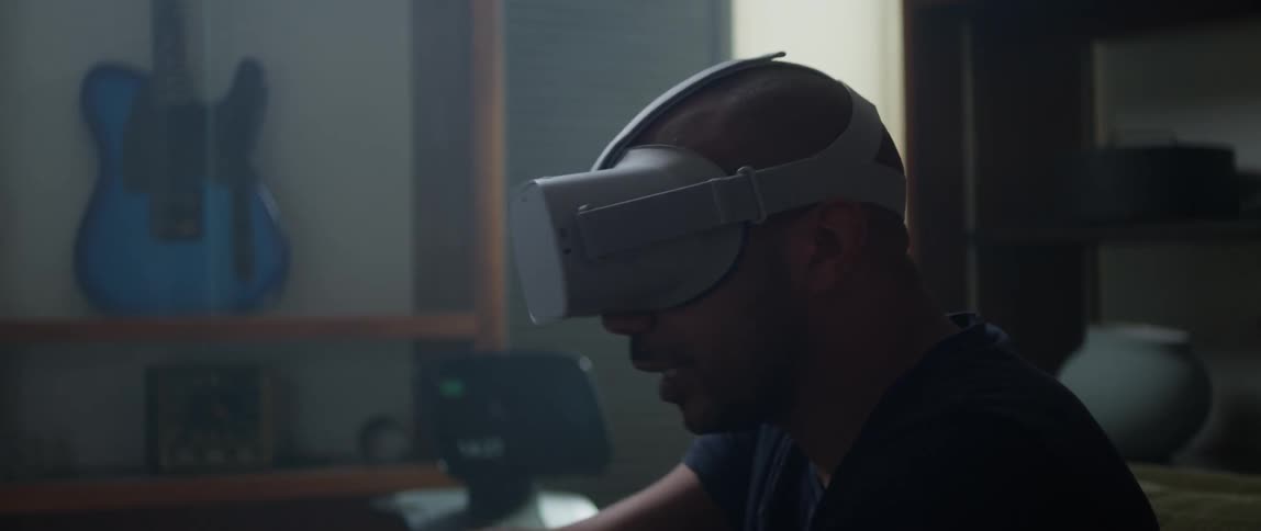 A man is sitting on a living room couch wearing a virtual reality headset and talking as he moves the controller