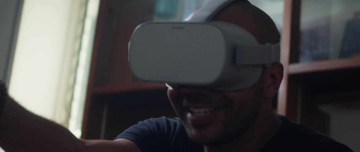 A man is sitting on the couch wearing a virtual reality headset smiles as he ducks and dodges