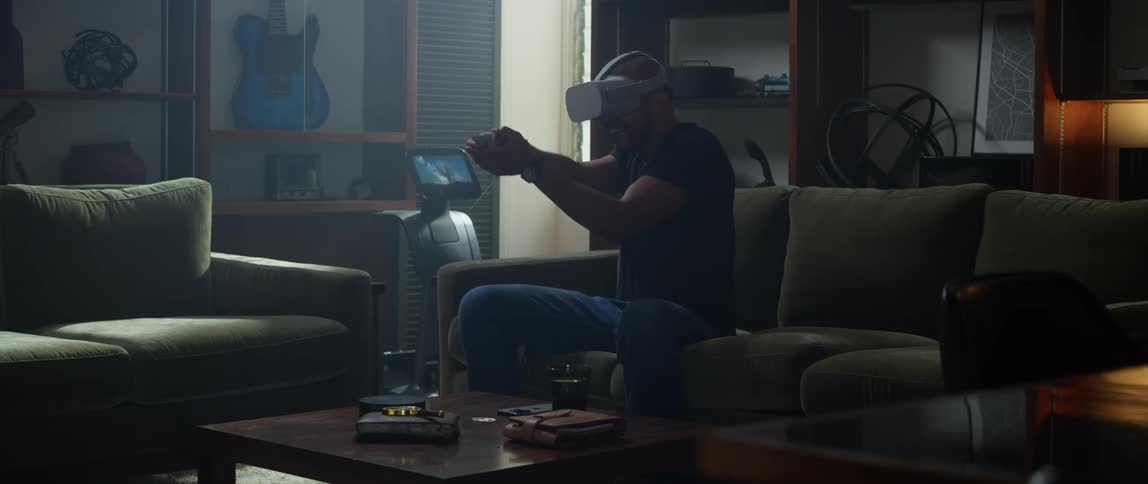 A man is sitting on a living room couch wearing a virtual reality headset and moving his arms with a remote controller as he plays