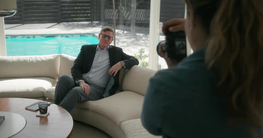 A man seated on a couch poses for a female photographer during a photo shoot