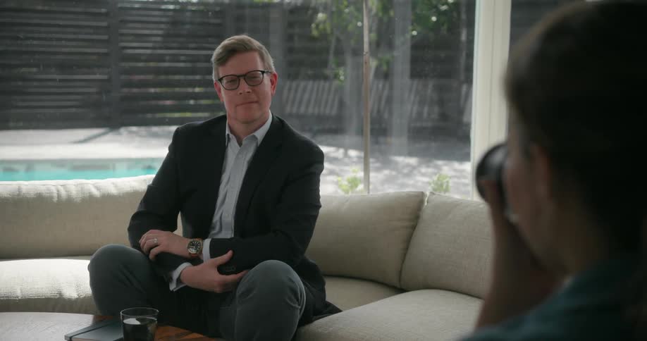 Man wearing black blazer jacket and pants sits on brown couch posing for the camera in living room at home