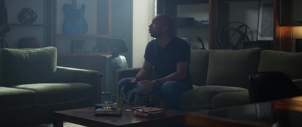 A man sits at home on a couch with virtual reality goggles on and acts like he is shooting a gun and then looks defeated and takes the goggles off and walks away