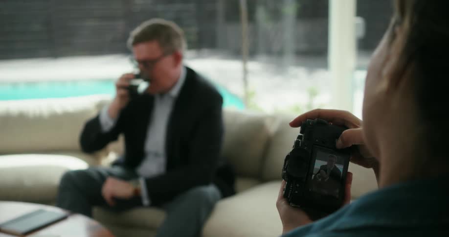 A man sits on a couch at home and poses while a woman takes a picture of him and then he picks up a glass and takes a drink