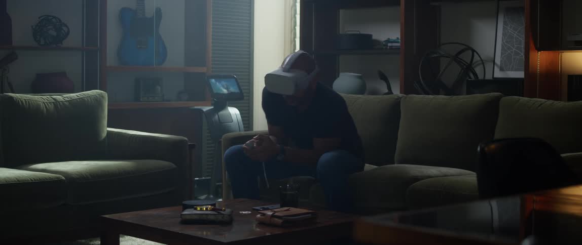 A man wearing a virtual reality headset sits on his couch and excitedly ducks and dodges objects in the game