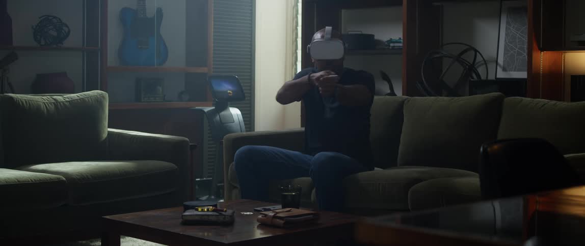 A man is sitting on a living room couch wearing a virtual reality headset and moving his arms with the remote controllers as he plays the game