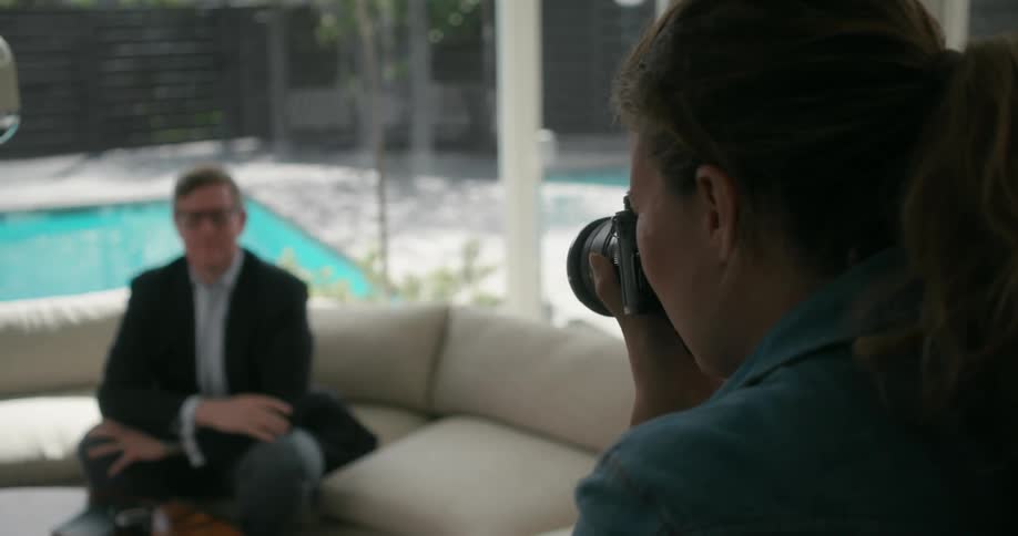 Female photographer takes pictures of man posing on couch with backyard pool in the background