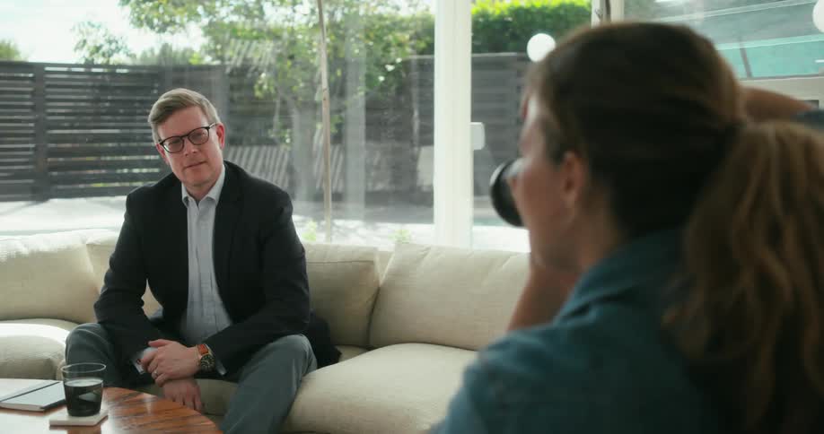 Female photographer takes photo of man wearing glasses blazer jacket and pants sitting on beige couch in the living room