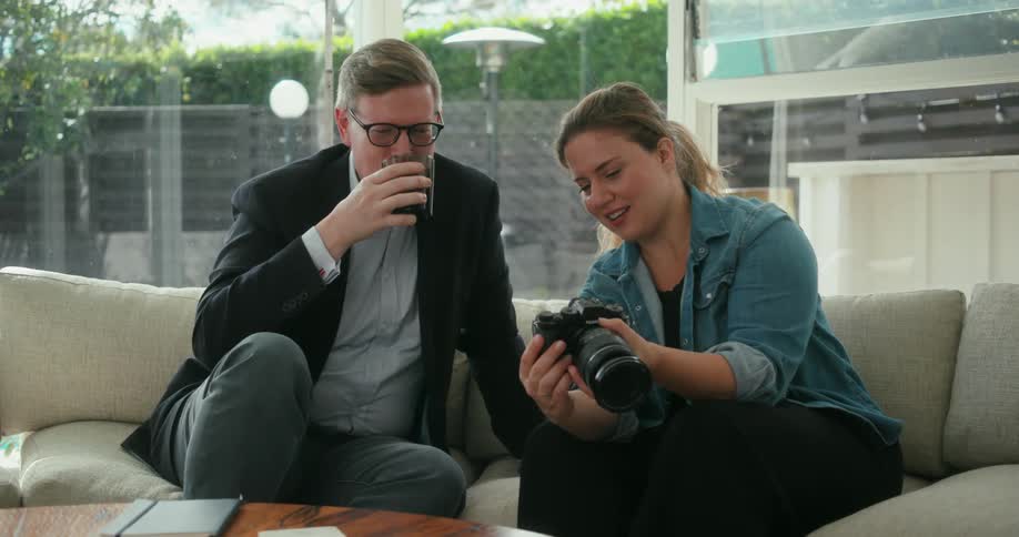 Man wearing glasses and blazer jacket shirt and pants drinks from glass looking at photos on camera with female photographer with ponytail blue shirt and black pants sitting on couch in living room