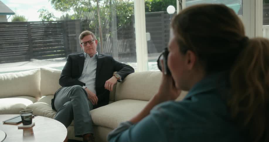 A man sits on a couch at home and poses for a photograph while a woman holding the camera instructs him how to move