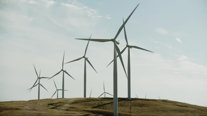 Wind turbines work on wind farm on hill with a vast blue sky filled with wispy clouds in the background as the dynamic shot tilts down