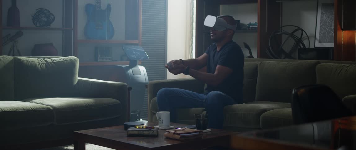 A man wearing a virtual reality headset sits on his couch while aiming his controller