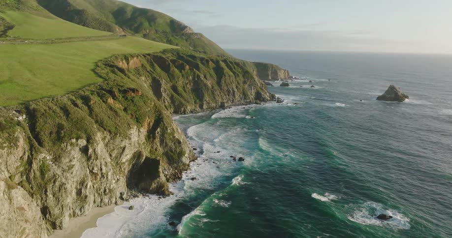 Waves crash against beach at base of cliff as traffic moves along nearby highway