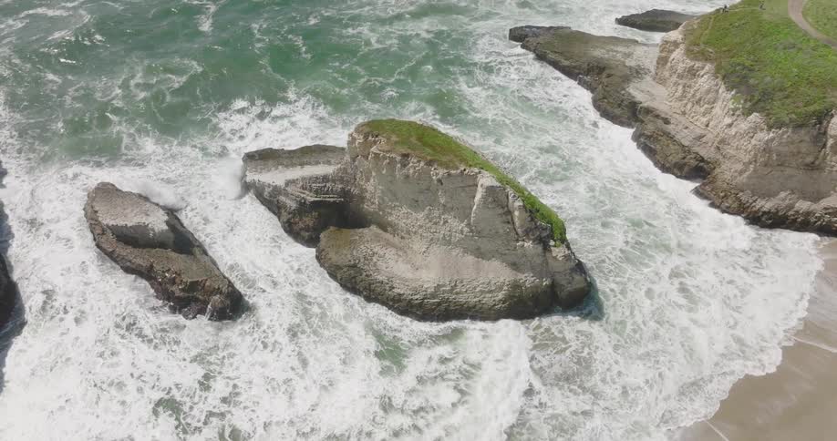 Aerial view of ocean waves breaking against big rock and coast at bay